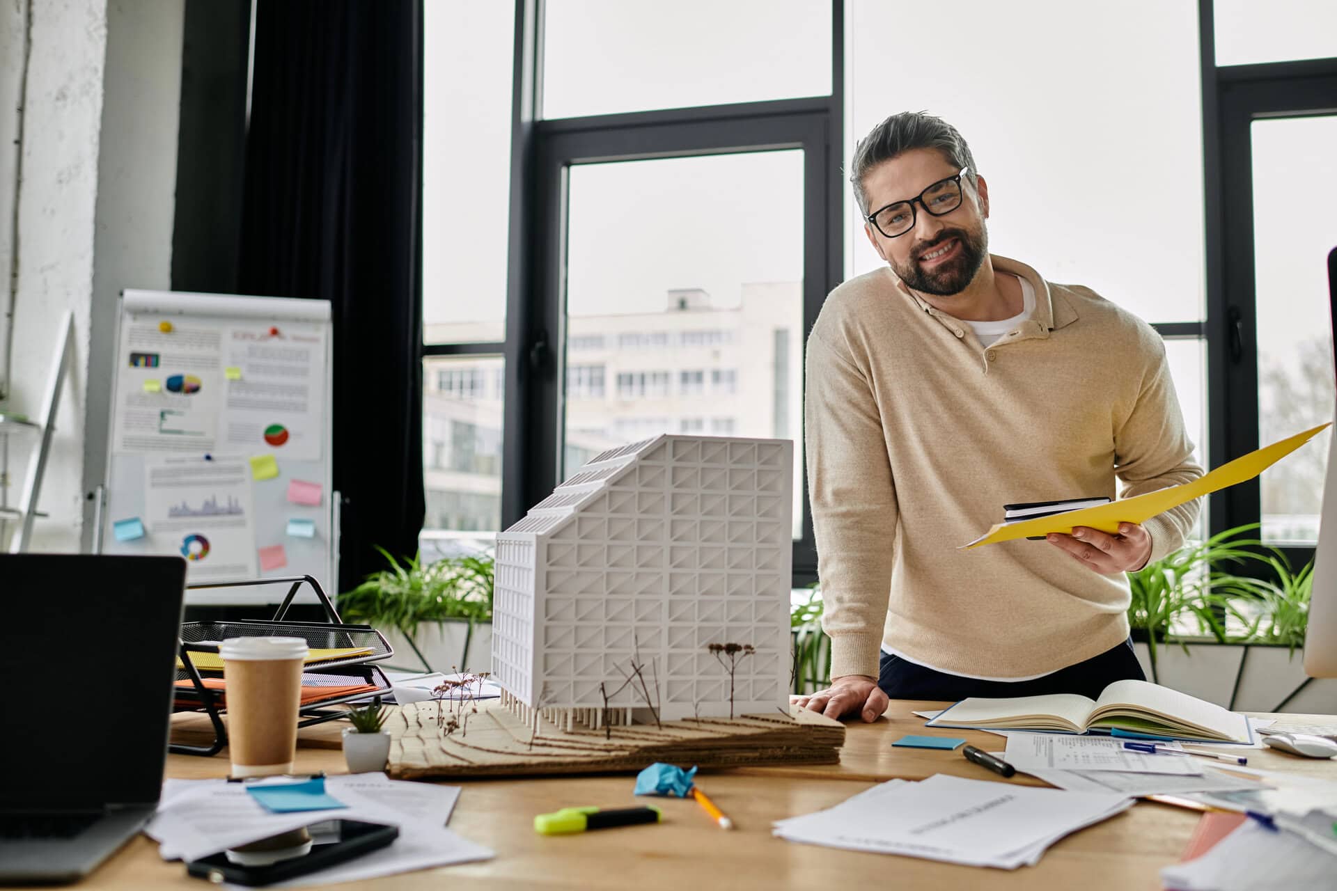 A handsome businessman with a beard reviews blueprints and a model building in a modern office.